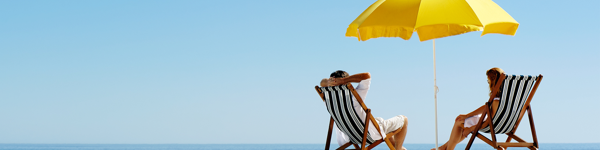 Couple sitting in beach chairs beside eachother with yellow umbrella in between them and wave in the background