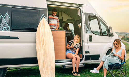 Three people relaxing around RV parked in campground.