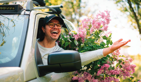 Man smiling and sticking arm out of car window with bouquets of flowers behind him in backseat.