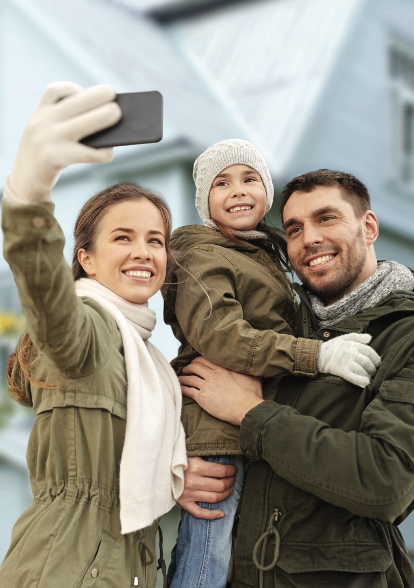 A happy family of three, dressed warmly in jackets, poses for a selfie outdoors. The parents smile while holding their child, conveying warmth and joy.