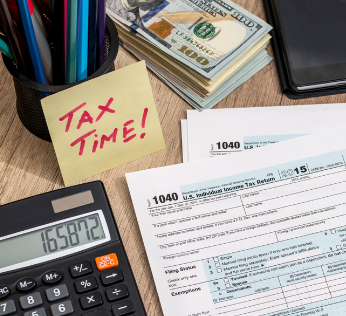 Alt text: "Tax-related items on a desk include IRS 1040 forms, a calculator showing 165872, a sticky note reading 'Tax Time!', and a stack of hundred-dollar bills."