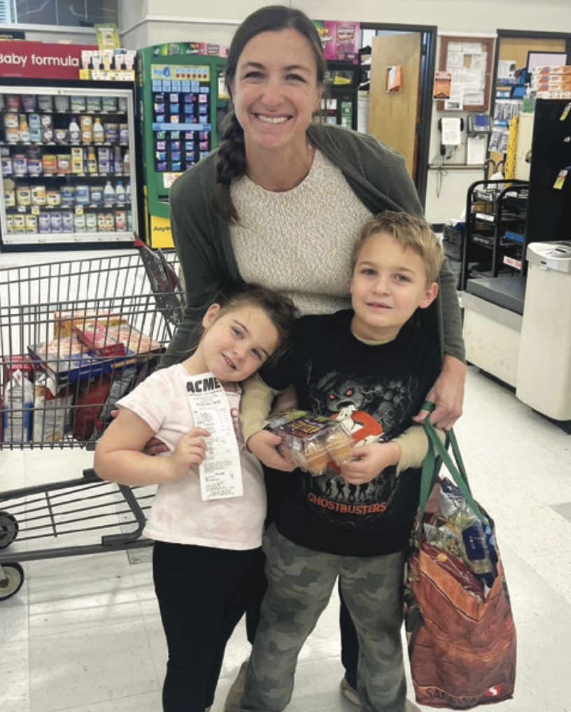 A smiling woman with two children stands in a grocery store. The girl holds a receipt and leans against the woman. The boy holds groceries. Shopping cart and shelves are in the background.