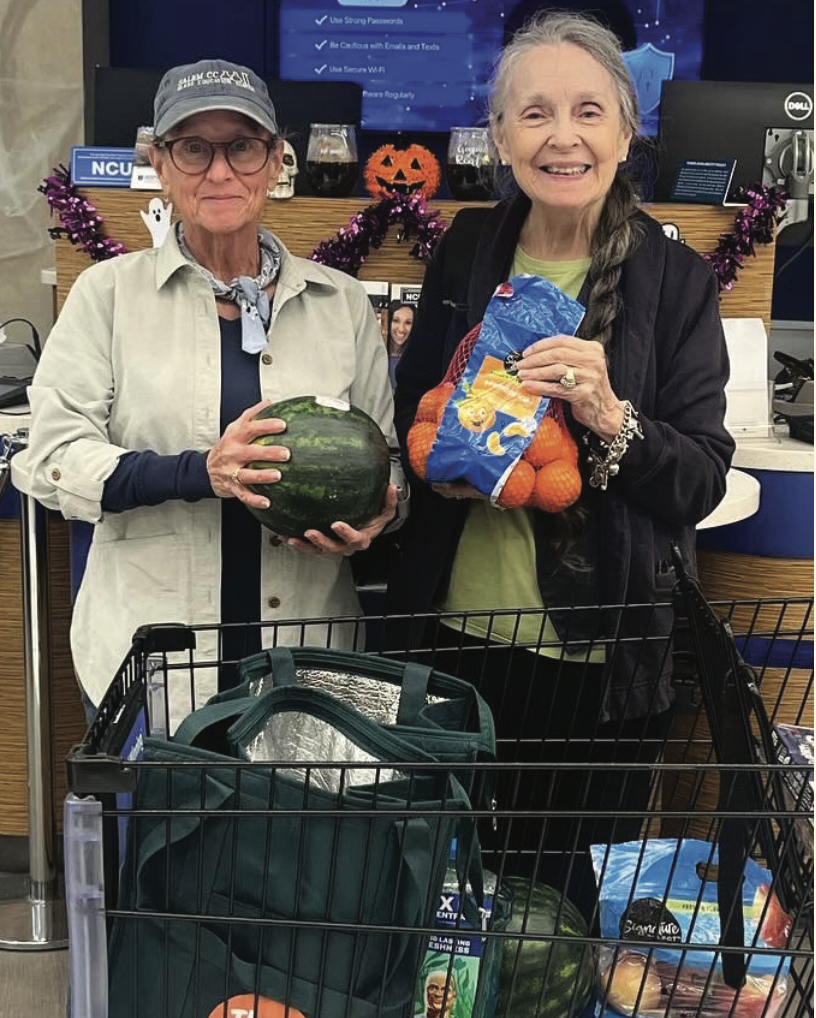 Two women stand smiling in a store. One holds a watermelon, the other a bag of oranges. A shopping cart and Halloween decorations are visible.