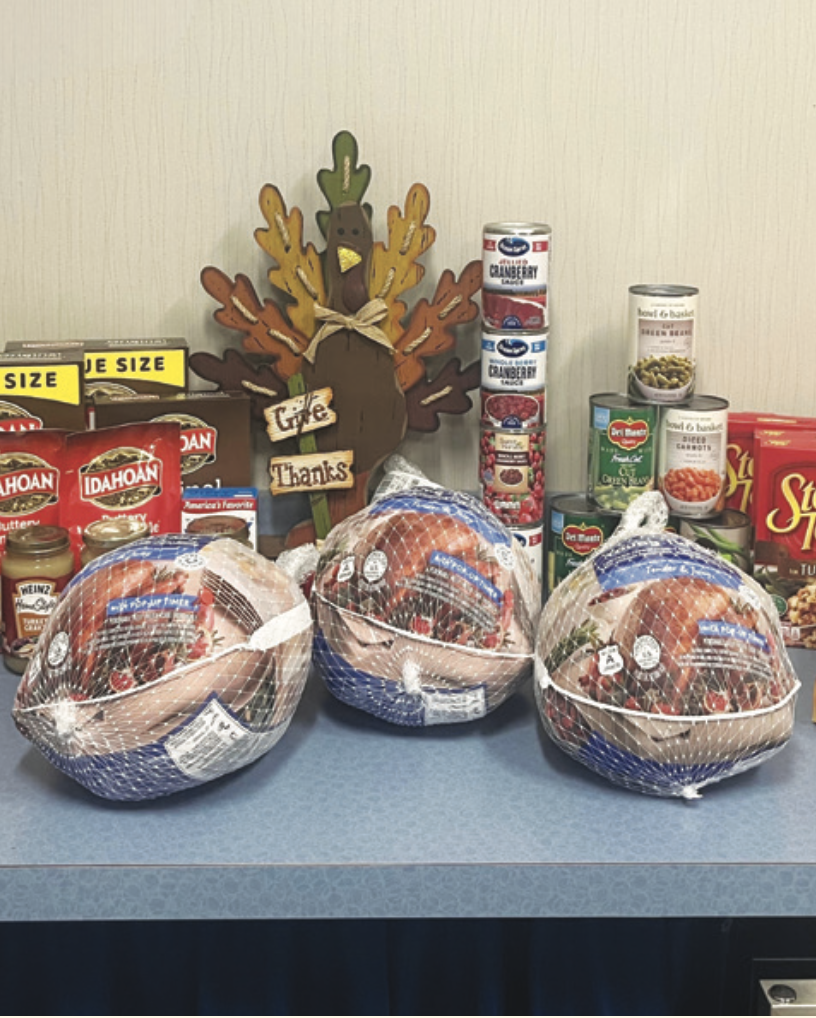 Three frozen turkeys sit on a table with canned vegetables, cranberry sauce, and instant potatoes. A wooden turkey decor with "Give Thanks" adds a festive touch.