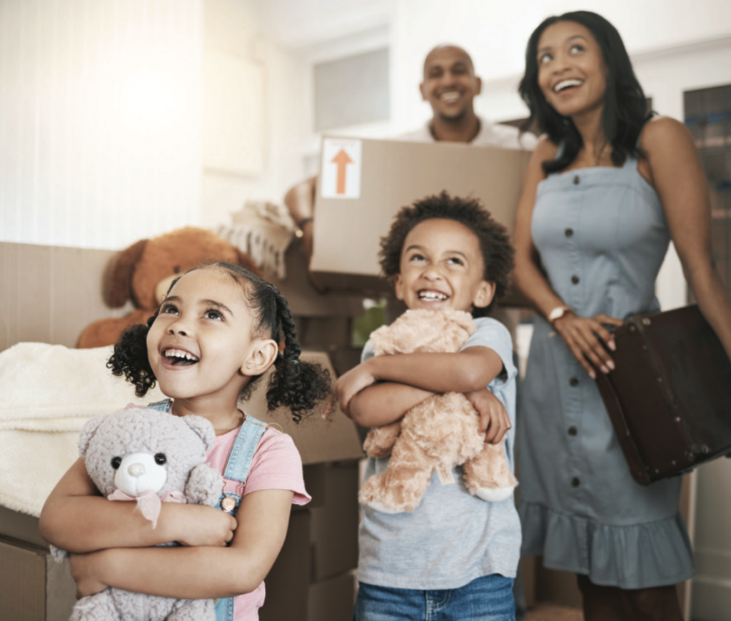 A joyful family of four in a new home with cardboard boxes. Two children smile, hugging teddy bears, while parents look on happily in casual clothes.